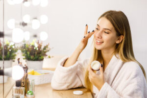 A woman in a white robe holds a skincare jar while applying product in front of a vanity mirror.