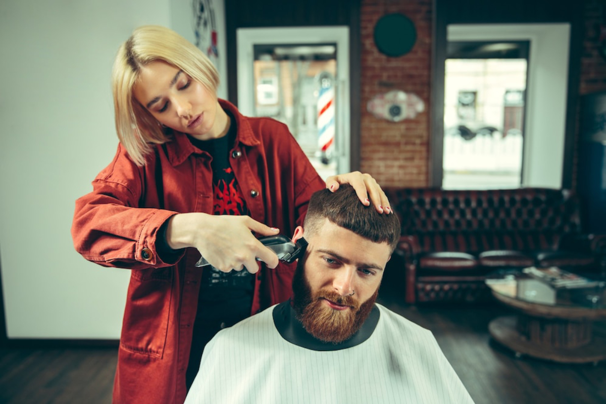 A hairstylist in a red jacket uses clippers to trim a client's hair in a stylish barbershop with a brick wall backdrop.