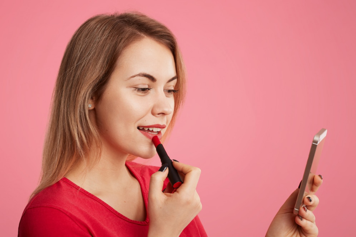 A woman with long blonde hair holds a lipstick in one hand and a smartphone in the other against a vibrant pink backdrop.