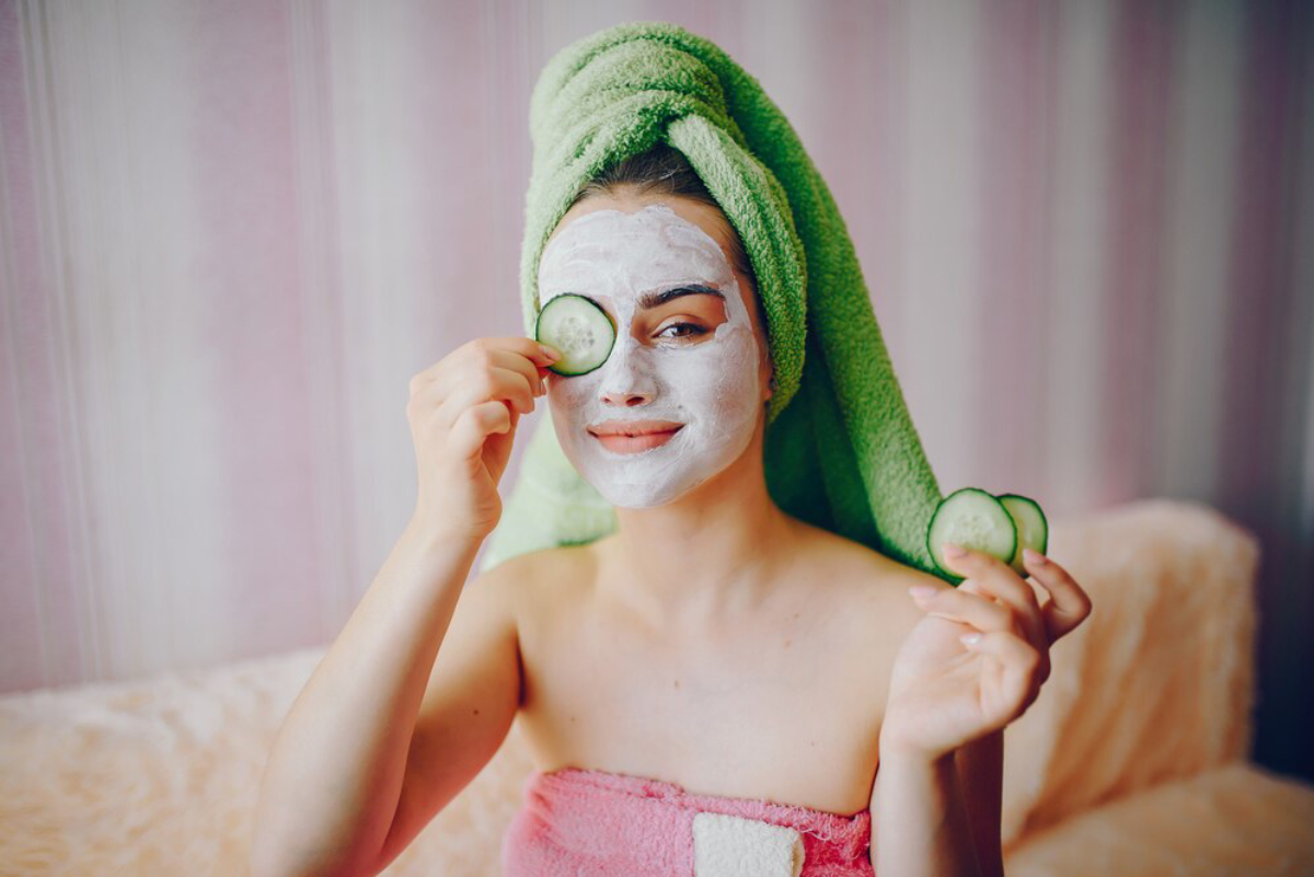 A person wearing a green towel on their head and a pink towel around their body holds a cucumber slice in one hand.