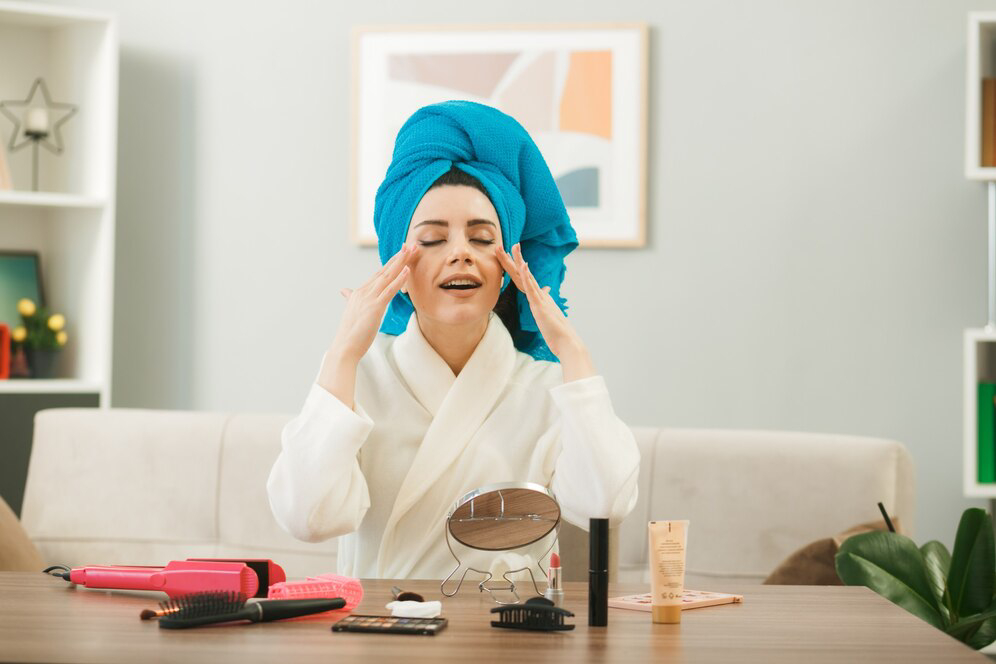 A person in a white robe and a blue towel gestures while seated at a table with beauty products and a mirror.
