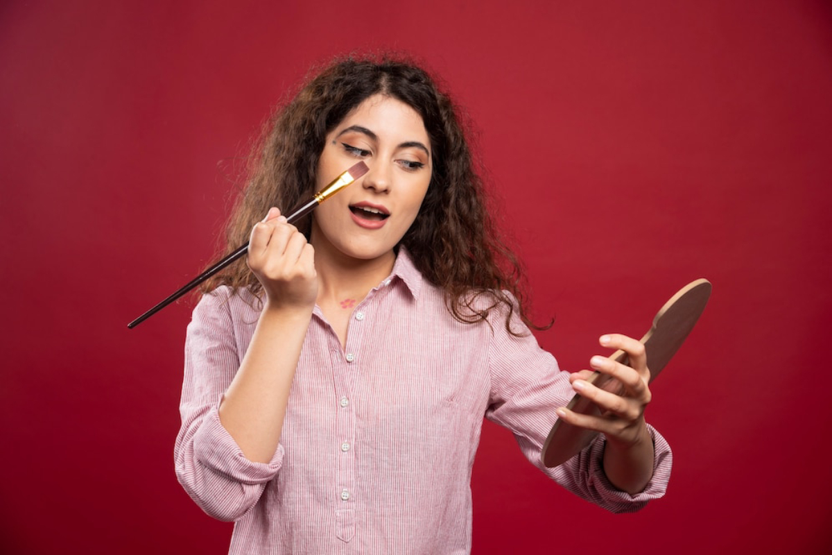A person with curly hair wears a collared pink shirt, holding a brush and a mirror against a vibrant red background.