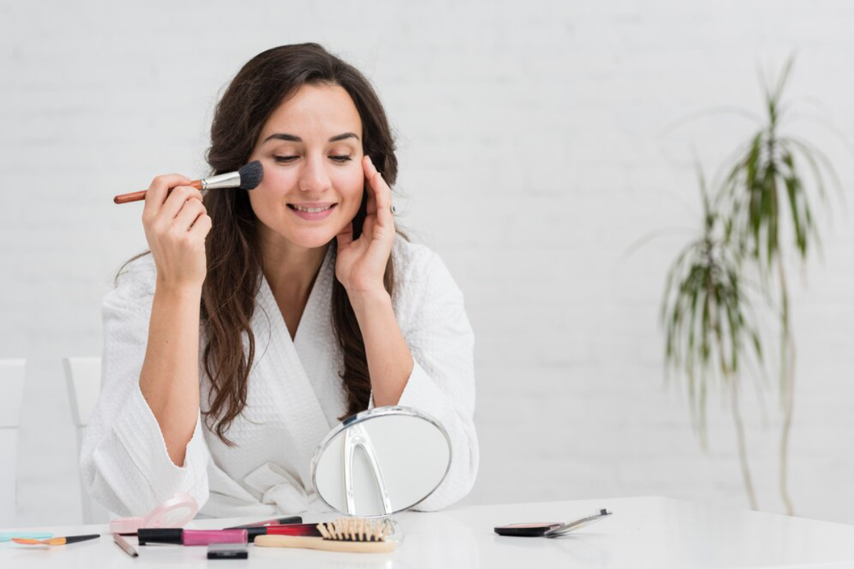 A woman in a white robe applies makeup in front of a mirror, surrounded by various cosmetics on a white table.