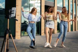 Three young women pose playfully outdoors, dressed in casual summer attire, beside a tripod with a smartphone.