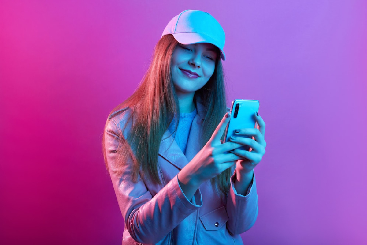 A person in a light pink jacket and blue cap, holding a smartphone.