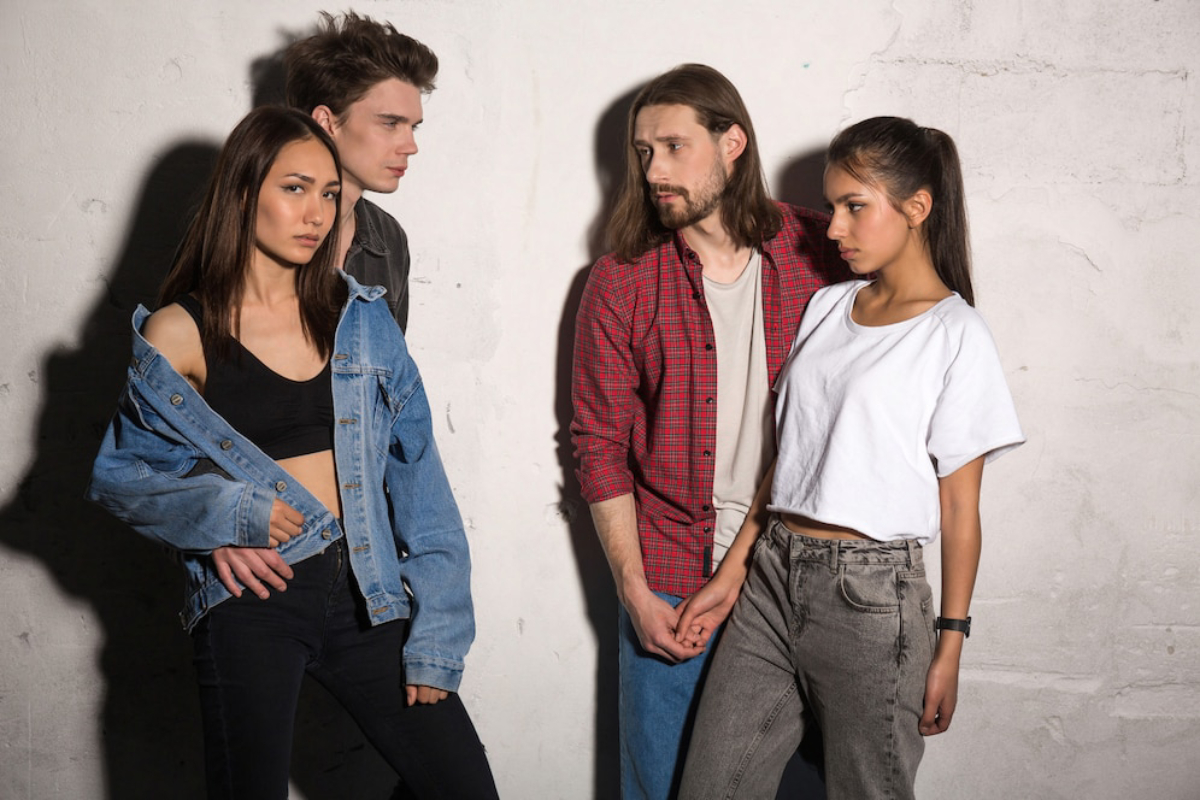 Four young adults dressed in casual attire pose against a textured wall, showcasing a blend of denim, plaid, and relaxed styles.