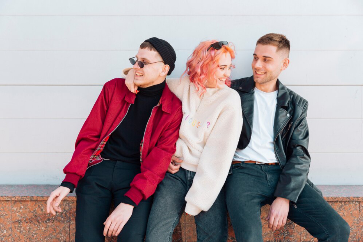 Three friends sit closely together on a low wall, casually dressed in colorful outfits against a light backdrop.