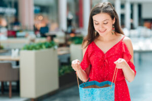 A woman in a red polka dot dress holds a blue shopping bag in a bright, modern mall setting.