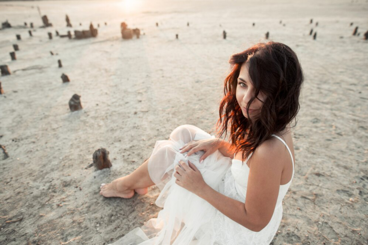 A woman in a white dress sits on a barren landscape at sunset, surrounded by wooden stumps and soft golden light.