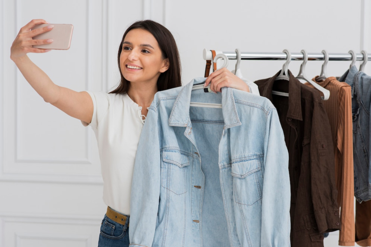 A woman holds up a light blue denim jacket while taking a selfie in front of a clothing rack displaying various garments.