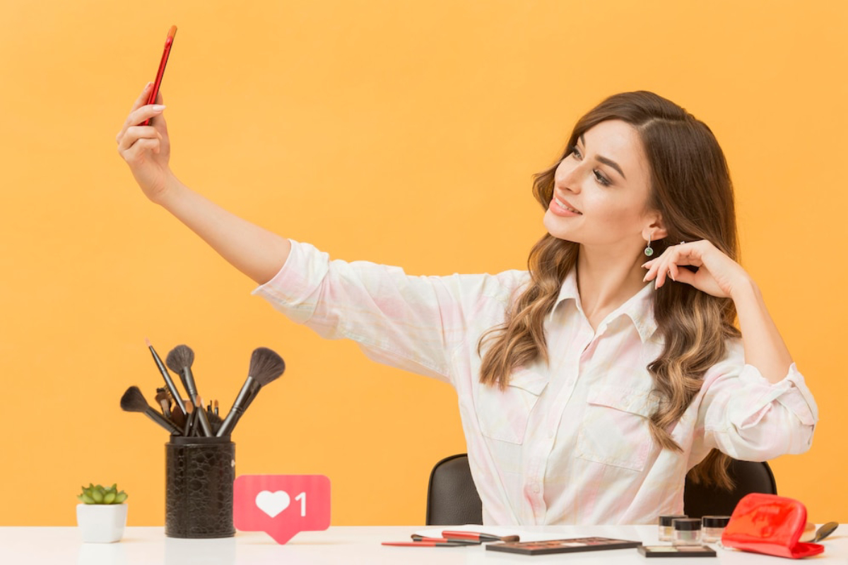 A person takes a selfie with a makeup brush in hand, surrounded by makeup tools.