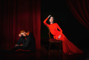A man in a black outfit sits on the floor, while a woman in a flowing red dress poses elegantly on a chair against dark curtains.