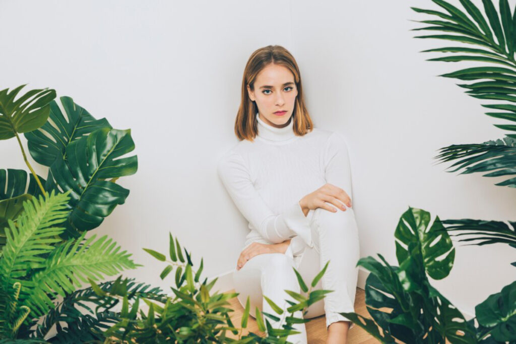 A person in a white turtleneck and pants sits against a white wall, surrounded by lush green tropical plants.