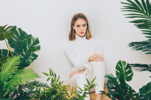 A person in a white turtleneck and pants sits against a white wall, surrounded by lush green tropical plants.