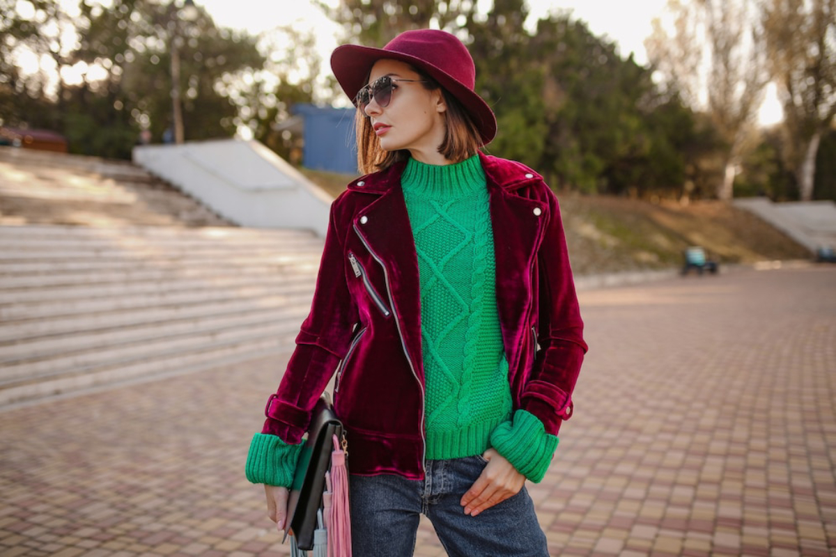 A person wearing a stylish burgundy velvet jacket, bright green sweater, and a hat, holding a bag in an outdoor setting.