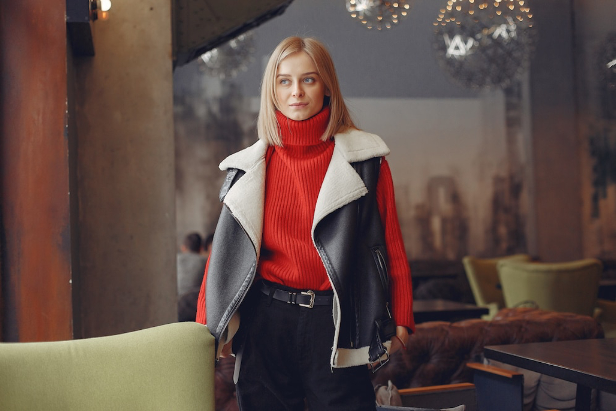 A person stands in a cozy café wearing a red ribbed sweater and a black and white shearling jacket, with stylish black pants.