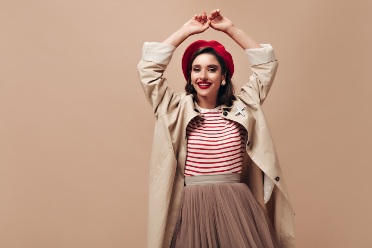 A woman poses happily in a beige trench coat, red beret, and striped shirt, with her arms raised against a neutral backdrop.