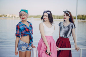 Three young women stand by a serene waterfront, showcasing vibrant outfits and playful hairstyles against a bright sky.
