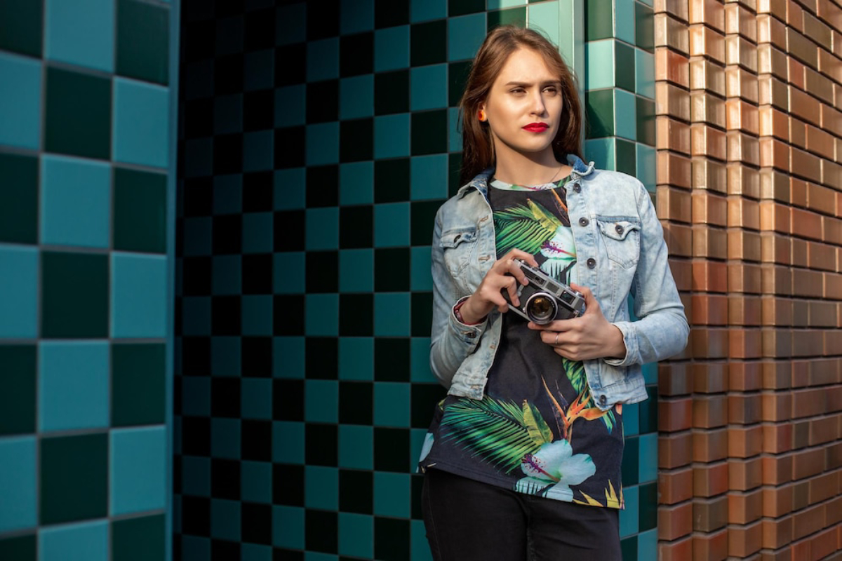 A person stands against a colorful tiled wall, holding a vintage camera, dressed in a floral shirt and denim jacket.