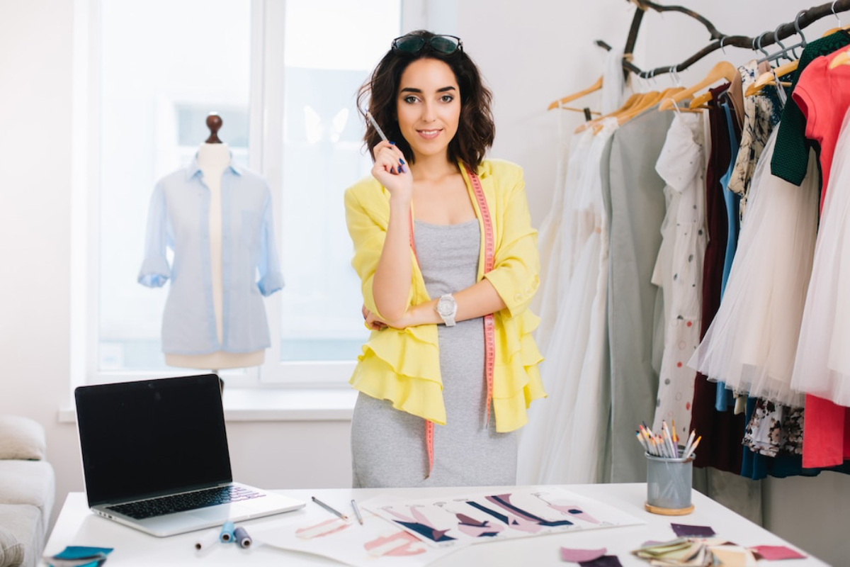 A fashion designer stands in a bright studio, surrounded by fabrics and sketches, pondering while holding a pencil.