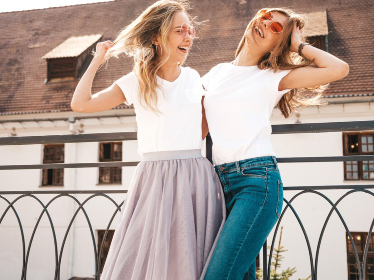 Two women are playfully posing outdoors, one in a white t-shirt and a gray skirt, the other in a white t-shirt and blue jeans.