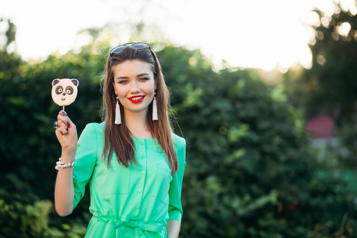 A young woman in a green shirt holds a panda-themed ice cream popsicle against a backdrop of lush greenery.