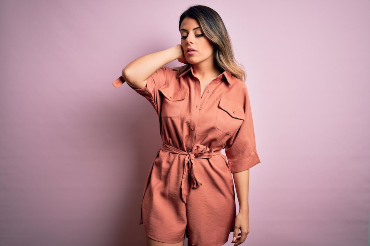 A person poses in a stylish, rust-colored shirt dress with a waist tie, against a soft pink backdrop.
