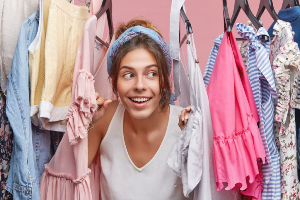 A person holds a pink and a gray shirt, surrounded by various colorful clothes on hangers against a soft pink background.