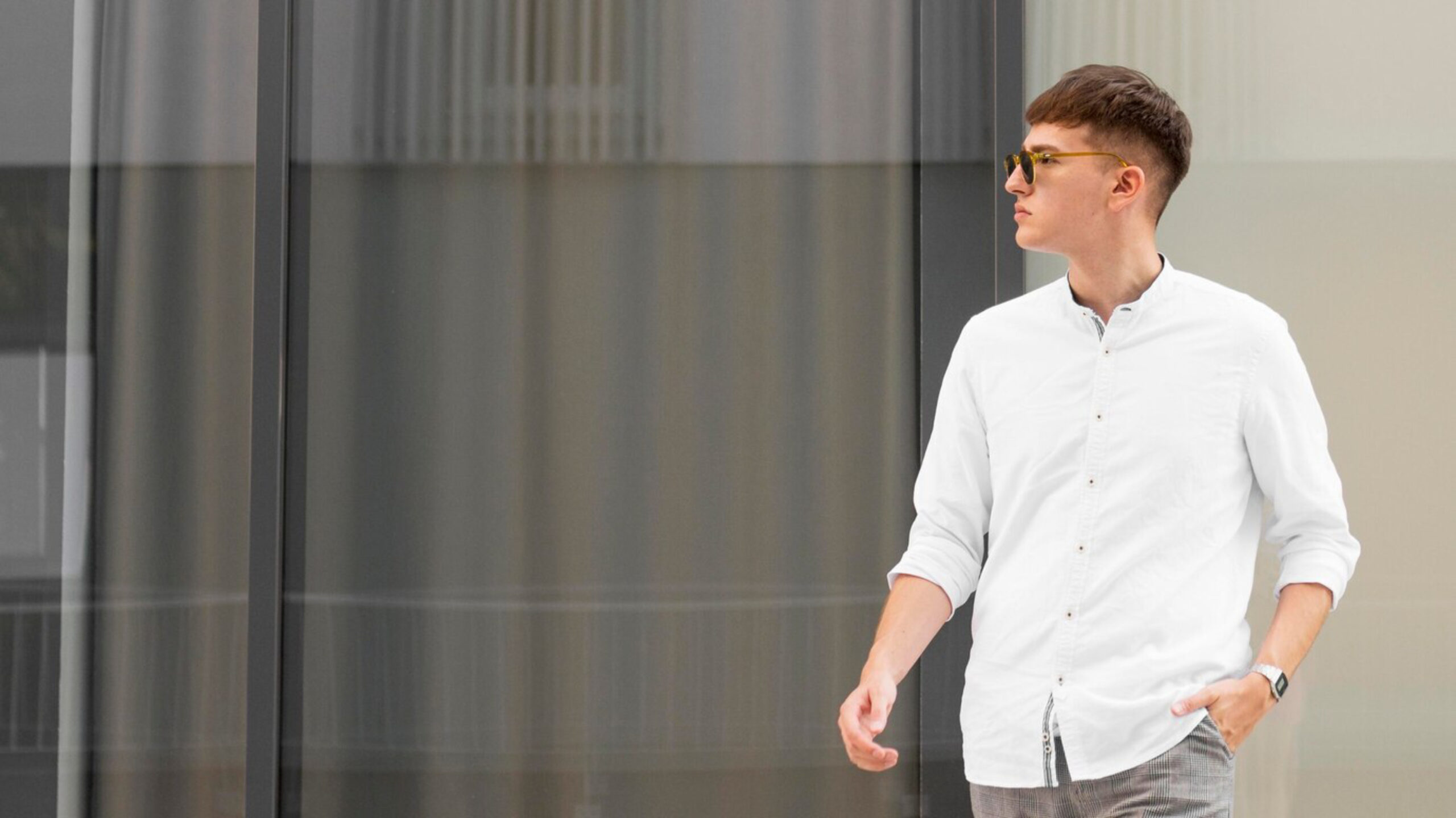 A man in a white button-up shirt walks casually past a modern glass facade.