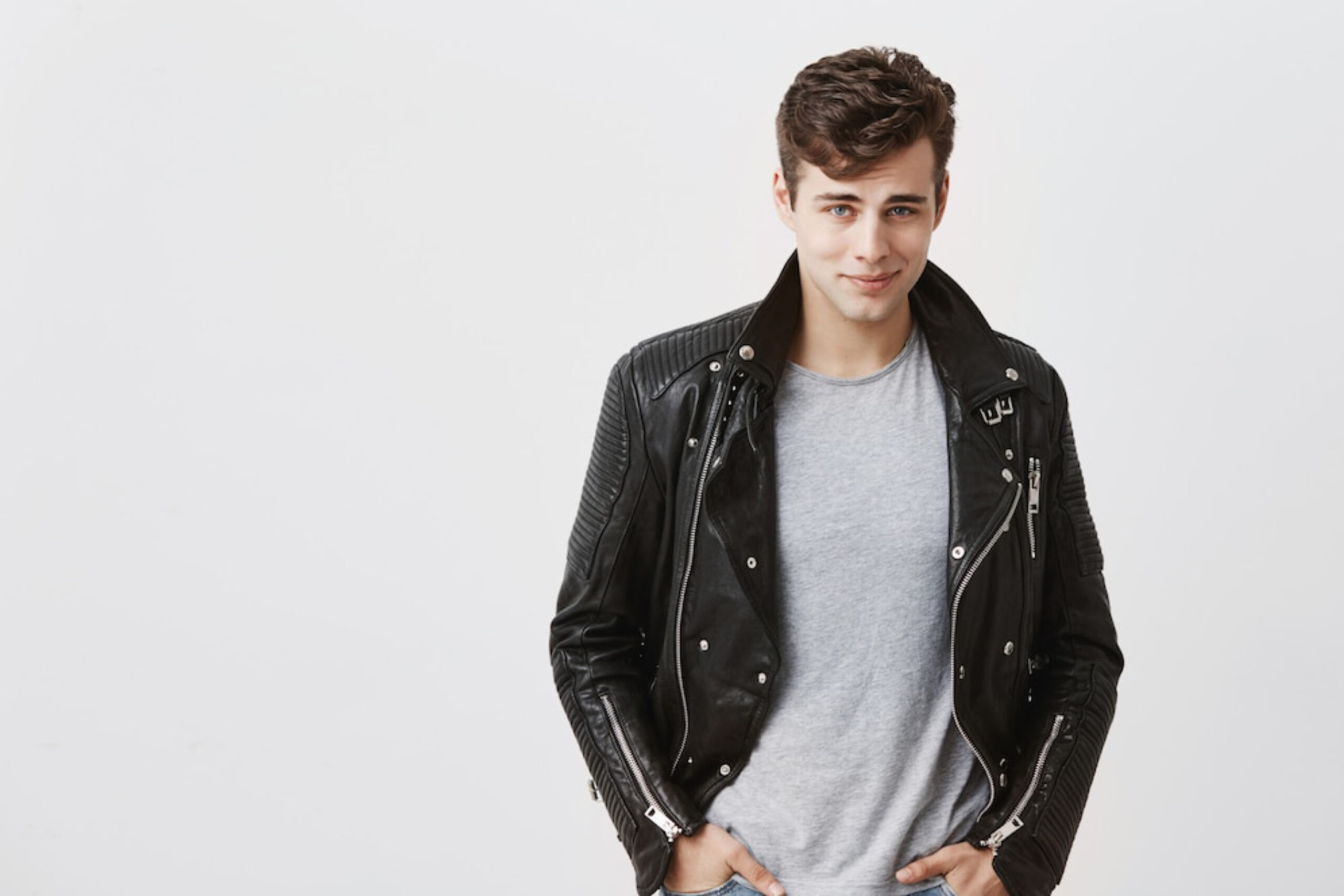 A young man wearing a black leather jacket over a gray t-shirt stands confidently against a plain white background.