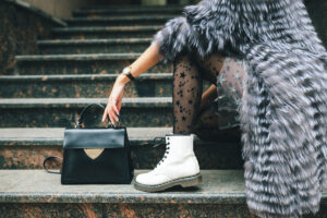 A fashionable woman in a furry coat sits on marble steps, showcasing star-patterned tights, white boots, and a chic black handbag.