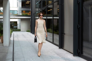 A woman in a form-fitting beige dress walks confidently down a modern stone pathway.