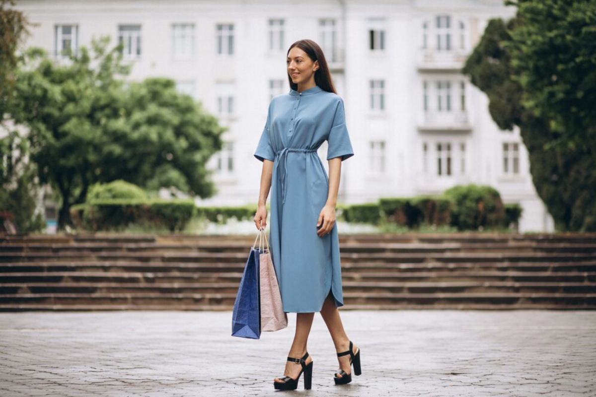 A woman in a stylish light blue dress stands on stone steps, holding two large shopping bags.