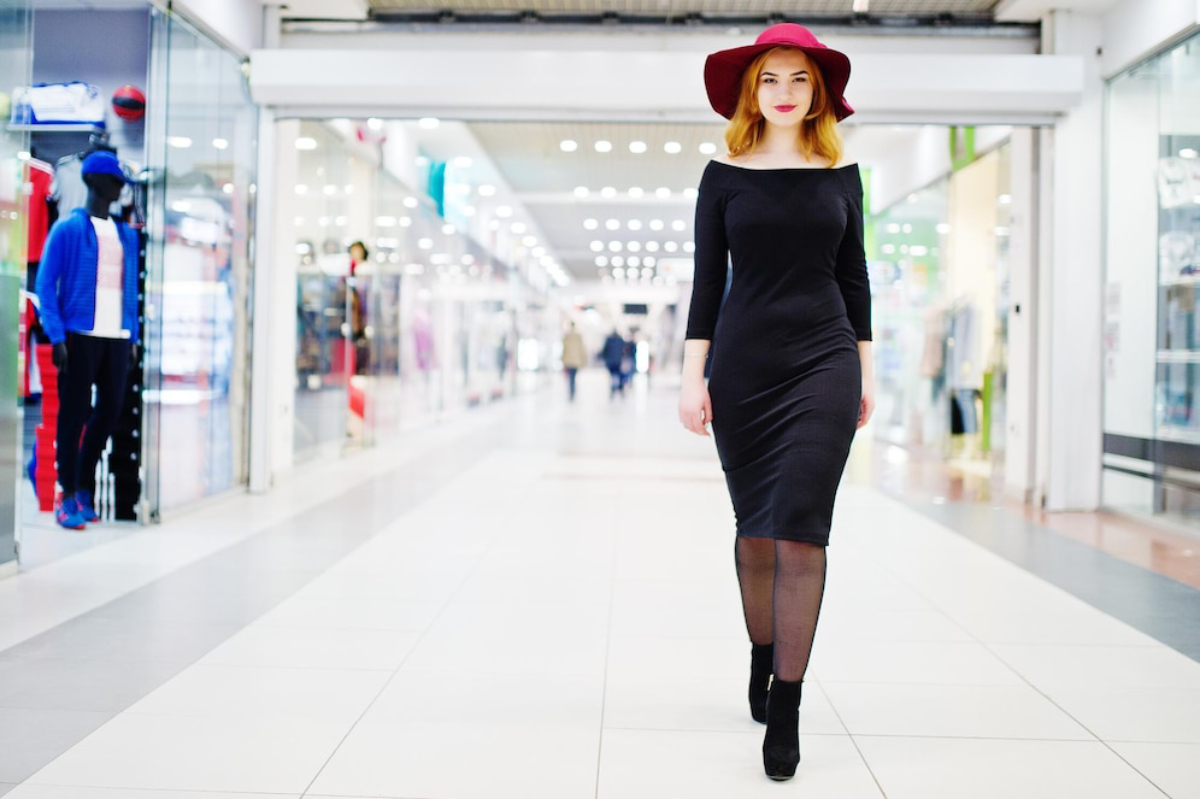 A woman walks confidently in a black off-shoulder dress and red hat, passing shops in a bright, modern shopping mall.