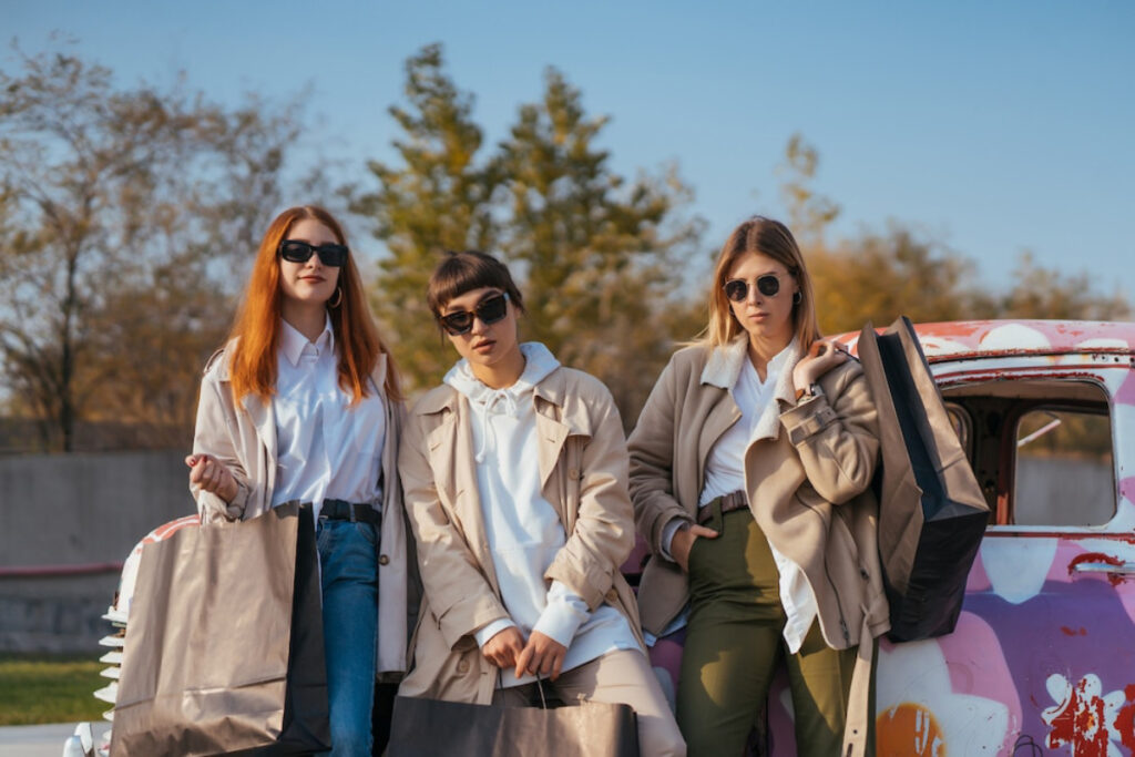 Three women in stylish outfits hold shopping bags, posed beside a vintage truck with colorful graffiti.