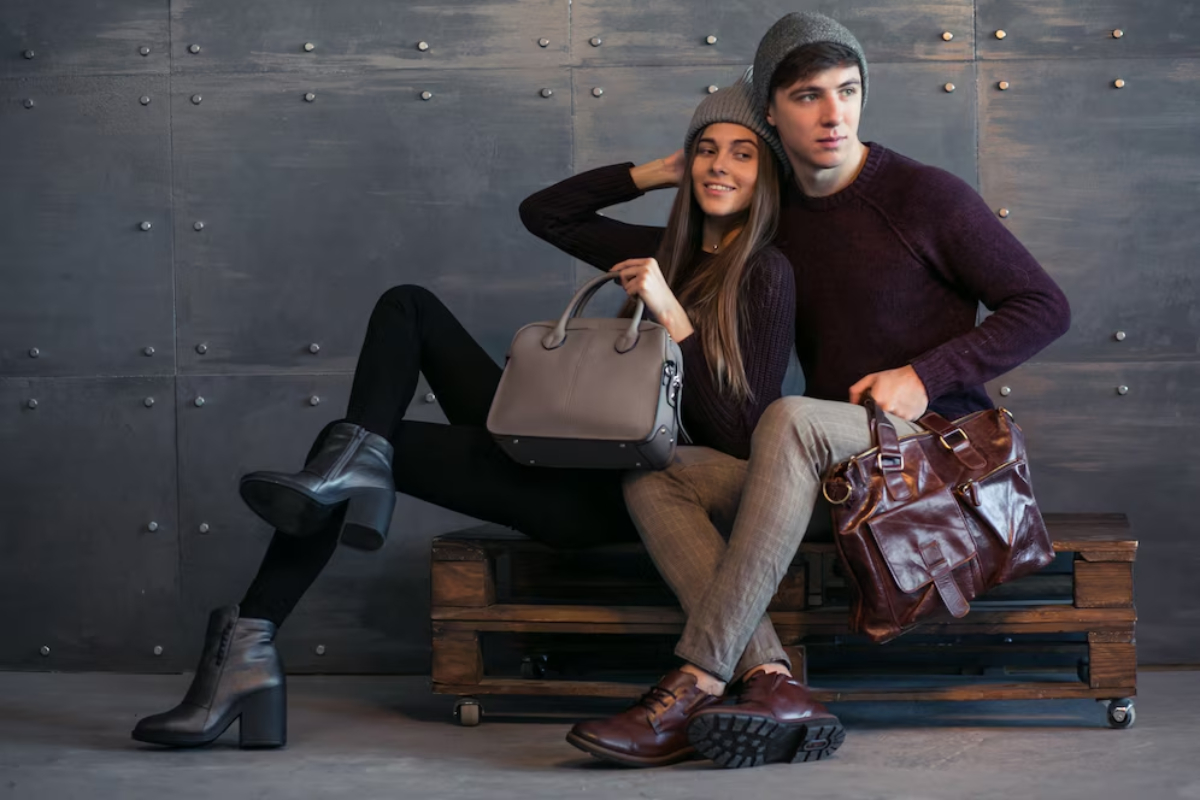 A stylish couple poses on a wooden bench, showcasing trendy outfits and fashionable bags against a textured metal backdrop.