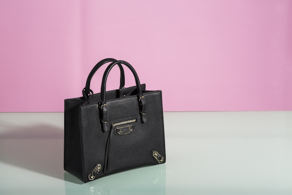 A stylish black handbag with structured design and gold accents, set against a soft pink background.
