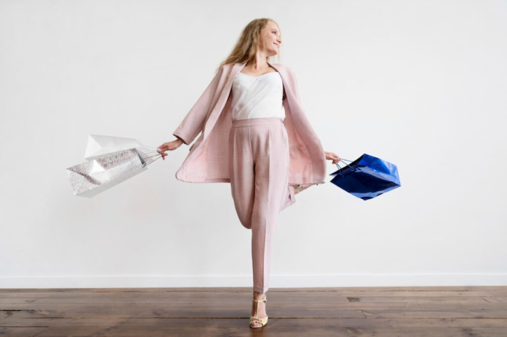 A stylish person wearing a pink suit strikes a pose while holding two colorful shopping bags against a clean.