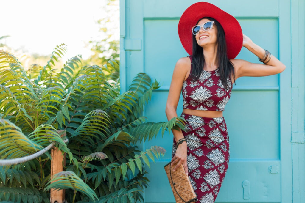 A woman in a maroon patterned two-piece outfit and a large red hat poses near lush green foliage and a teal background.