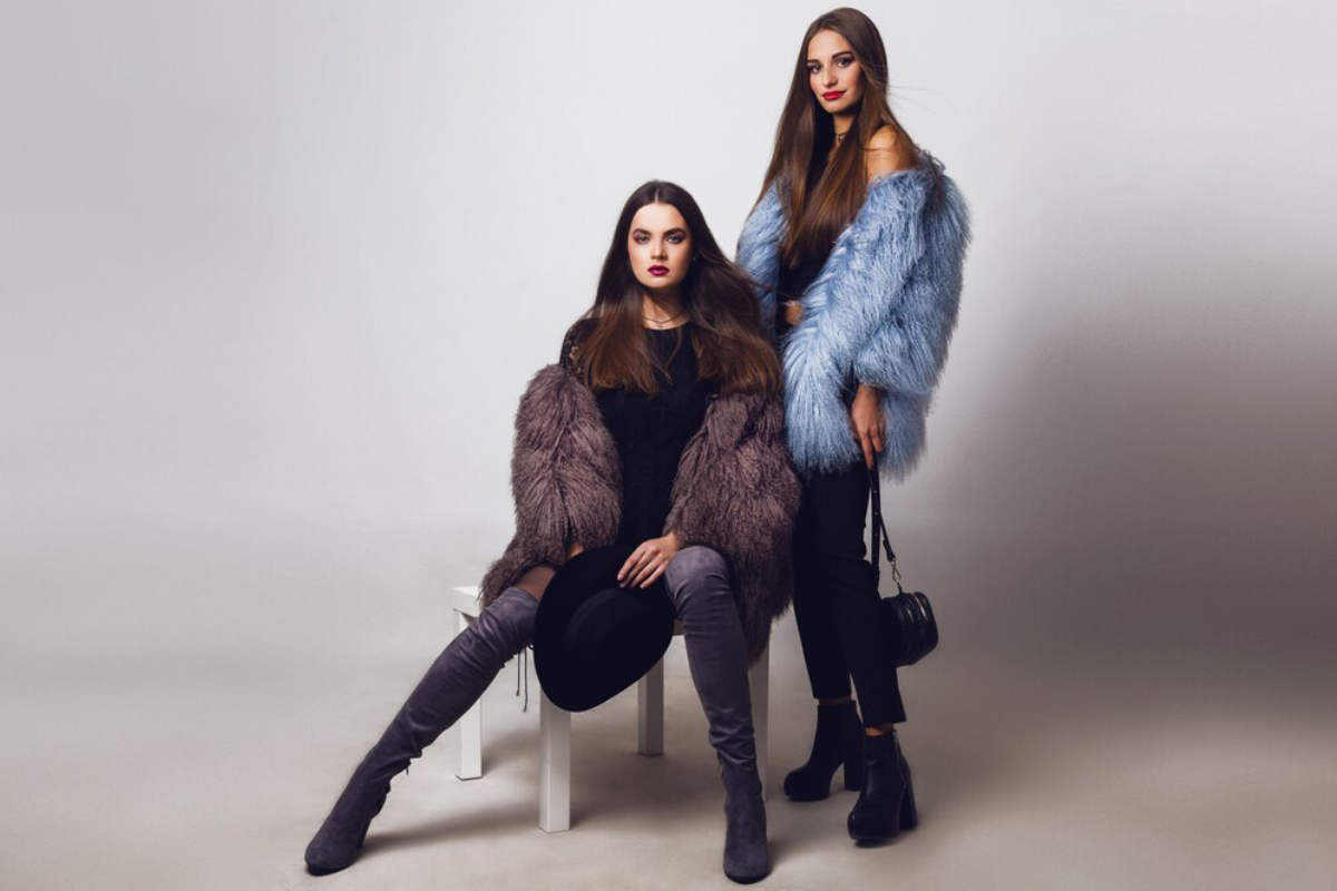 Two women pose stylishly in fluffy coats, one in purple and the other in blue, showcasing trendy outfits.