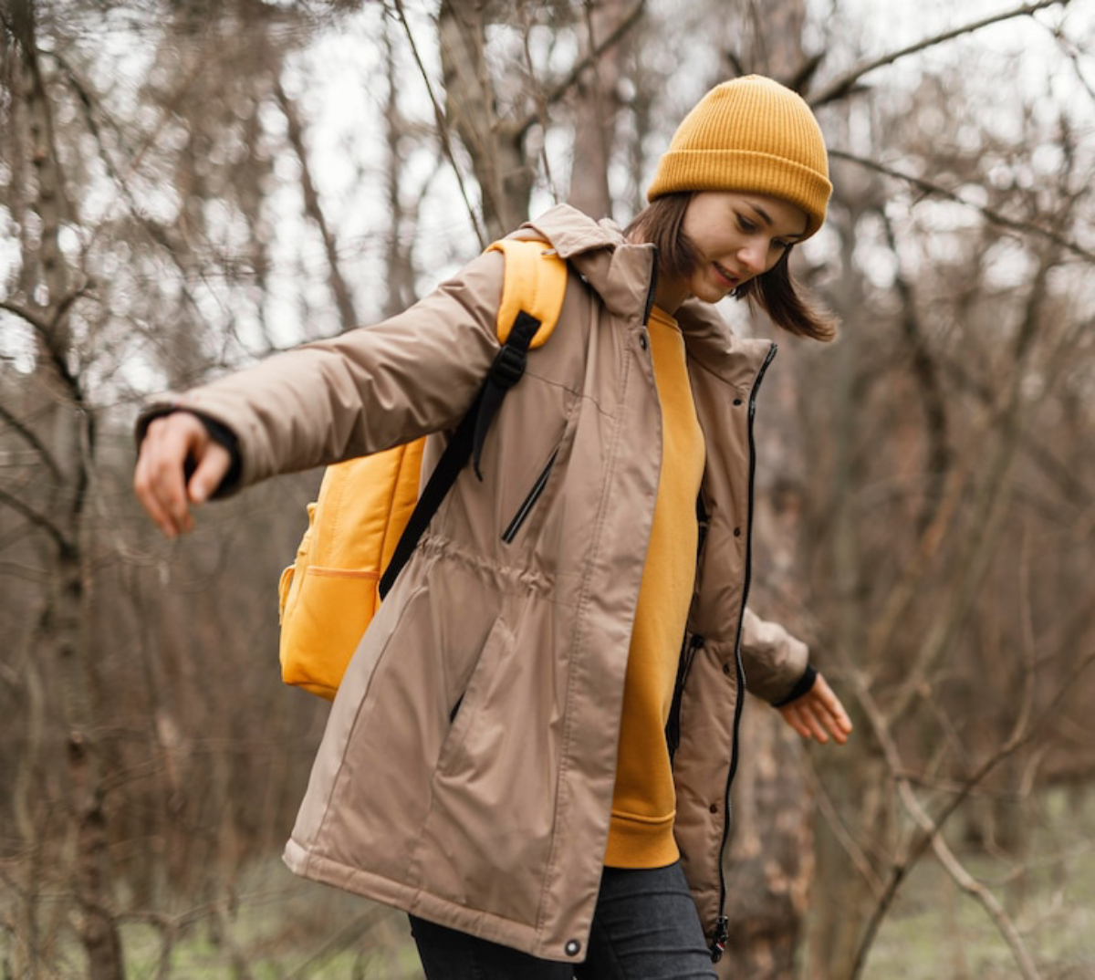 A person in a beige jacket and yellow beanie walks in a forest.