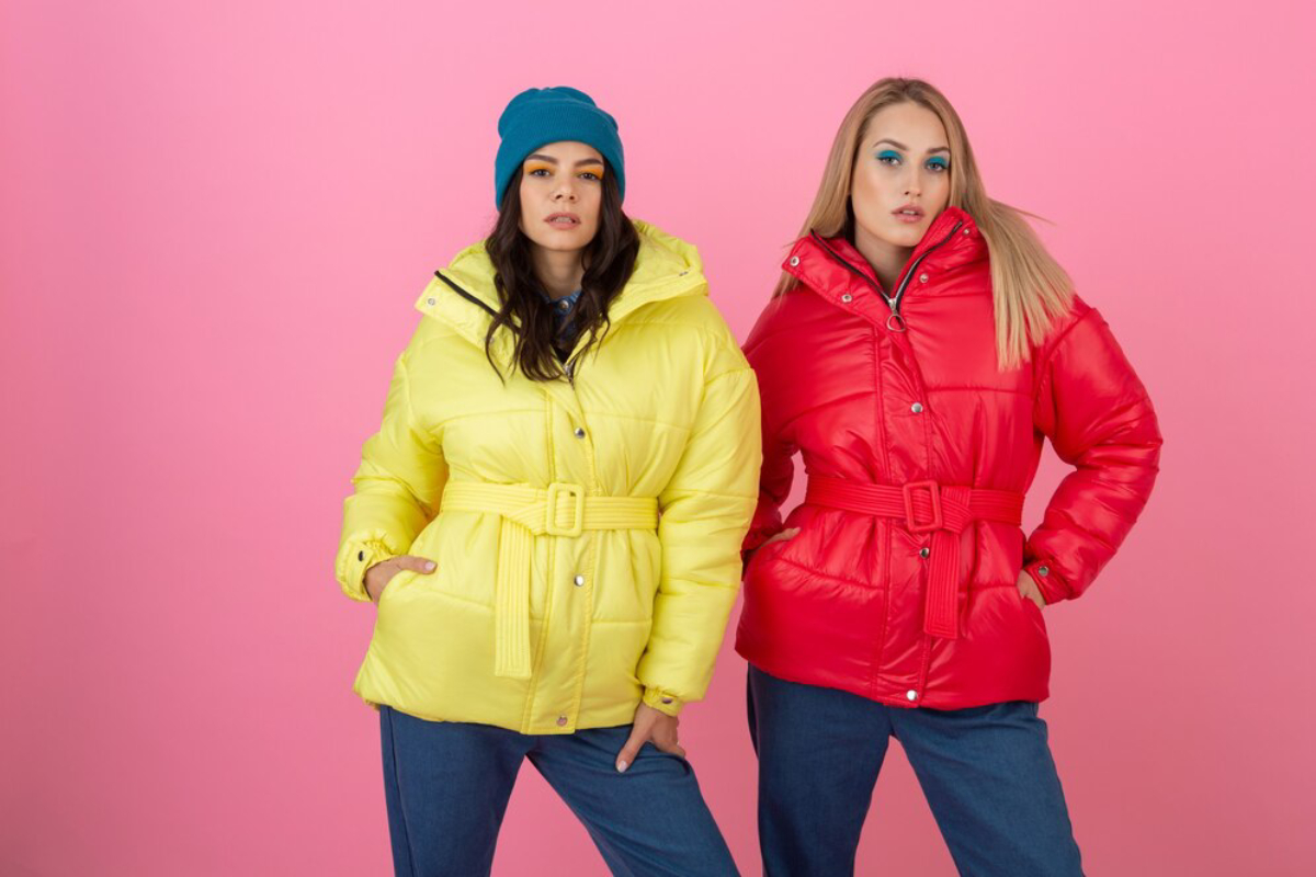 Two women stand confidently against a pink background, wearing vibrant yellow and red belted puffer jackets.