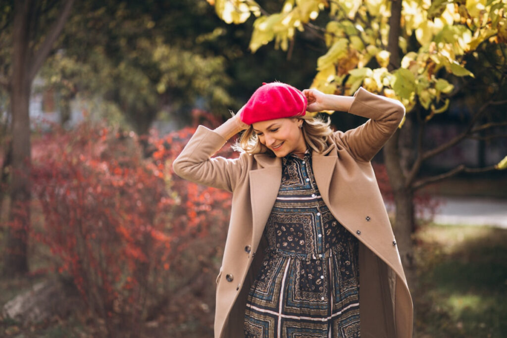 A woman in a stylish coat and vibrant pink hat poses playfully outdoors, surrounded by colorful autumn foliage.