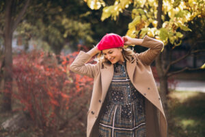 A woman in a stylish coat and vibrant pink hat poses playfully outdoors, surrounded by colorful autumn foliage.