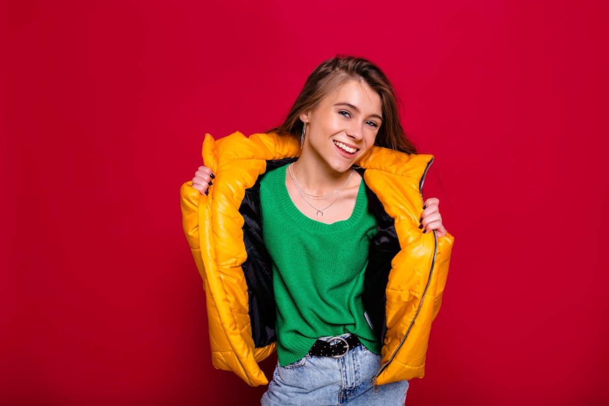 A person wears a vibrant yellow puffer jacket over a green sweater, set against a bold red background.