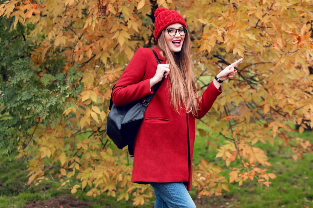 A woman in a red coat and matching hat stands against a backdrop of autumn foliage, gesturing with her hand.