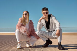 Two stylish individuals pose in an urban setting, dressed in trendy outfits against a clear blue sky.