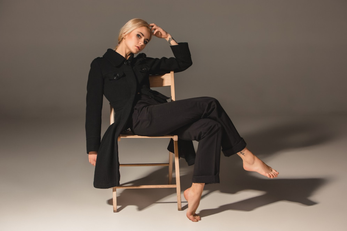 A person sits casually on a wooden chair, wearing a long black coat and black pants, with bare feet resting on the floor.