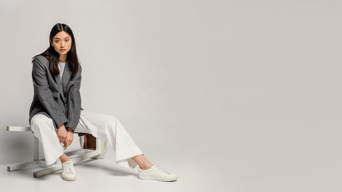 A model sits on a white stool, dressed in a grey blazer, white wide-leg pants, and minimalist white sneakers.