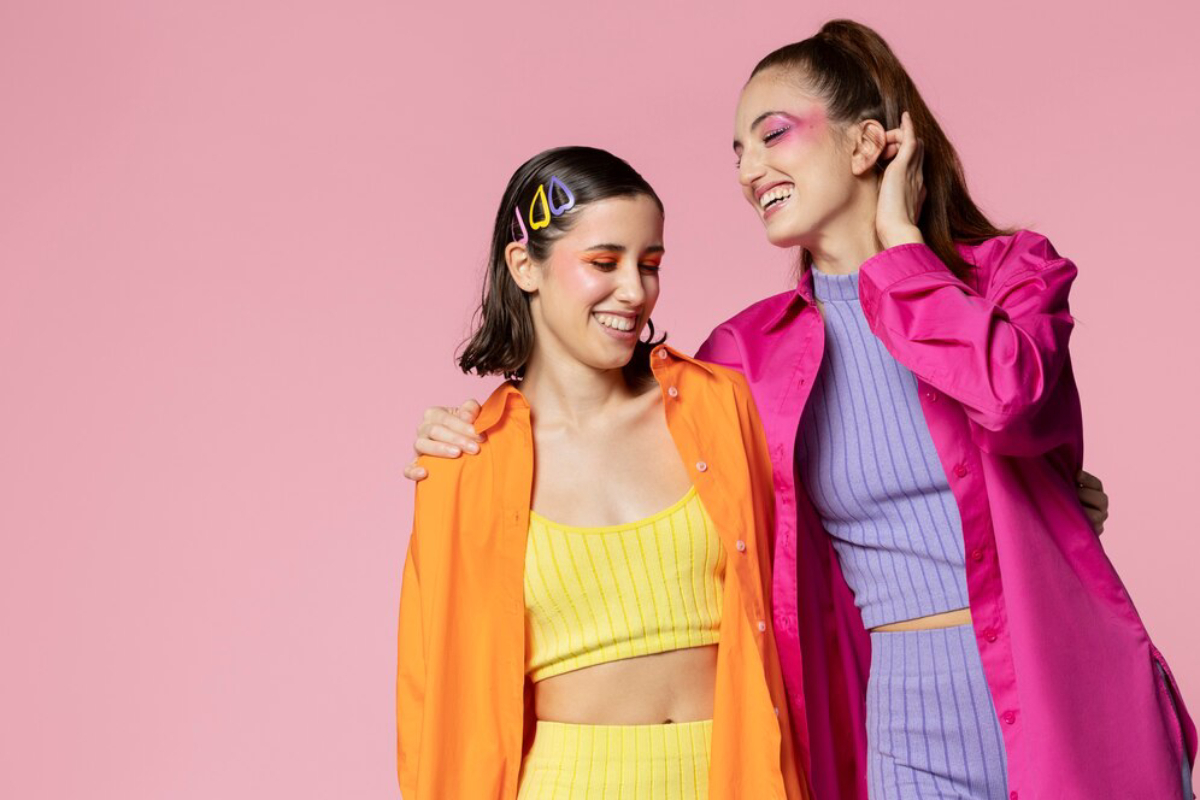 Two women pose against a pink background, wearing bright colored outfits—one in yellow and orange, the other in pink and purple.